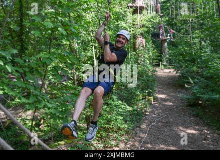 PRODUKTION - 19. Juli 2024, Hessen, Wiesbaden: Thorsten Held, Geschäftsführer von Kletterwald Neroberg, gleitet mit einer Seilbahn durch den Kurs. Der Kletterpark für junge und erfahrene Freizeitsportler liegt auf Wiesbadens Hausberg, umgeben von Buchen und Eichen. Der höchste Punkt ist ein Abenteuernest in einer Höhe von 25 Metern. Foto: Arne Dedert/dpa Stockfoto