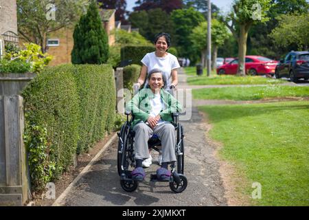 Alte indische Frau im Rollstuhl draußen auf einem Bürgersteig in einer Vorstadtstraße im Sommer, Großbritannien. Mutter und Tochter: Kann auch eine Betreuerin darstellen, die sich um die Ko kümmert Stockfoto