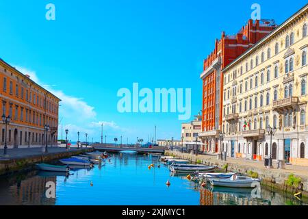 Boote fahren entlang eines malerischen Kanals, umgeben von üppigem Grün und historischen Gebäuden in Triest Italien. Wasserkanal mit einer Reihe von Booten und einer Brücke Stockfoto