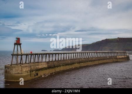 Ein langer Pier mit einem kleinen roten Leuchtturm am Ende, der sich unter einem bewölkten Himmel ins Meer erstreckt. Klippen sind im Hintergrund sichtbar. Stockfoto