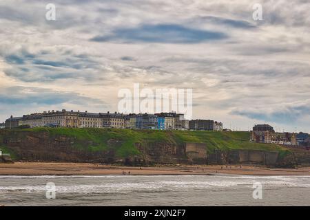 Eine Küstenstadt mit einer Reihe von Gebäuden auf einer Klippe mit Blick auf einen Sandstrand und das Meer. Der Himmel ist bewölkt und einige Leute laufen auf dem beac Stockfoto