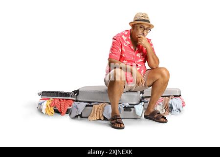Trauriger reifer männlicher Tourist, der auf einem vollgepackten Koffer mit unordentlichen Klamotten sitzt, isoliert auf weißem Hintergrund Stockfoto