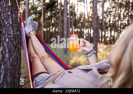 Frau, die sich in der Hängematte im Wald entspannt und Orangensaft oder Smoothie trinkt Stockfoto