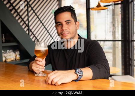 Stilvoller Latin-Mann in legeren Kleidern genießt ein Glas Bier an der Bar einer modernen Bar. Der Innenraum verfügt über einzigartige Beleuchtung und conte Stockfoto