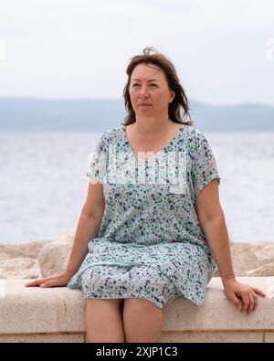 Glückliche Frau mittleren Alters sitzt am Meer und schaut nachdenklich in die Kamera. Reisen und neue Orte Stockfoto