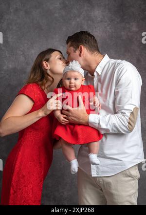 Ein sanfter Kuss junger Eltern, die ihre kleine Tochter im Arm halten. Europäische Familie und Kind. Das Mädchen und das Baby tragen rote Kleider Stockfoto