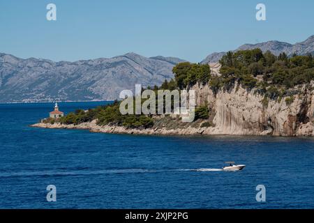 Ein kleines Boot fährt in den malerischen Hafen. Fantastische Küste. Die Berge sind mit Grün bedeckt, das blaue, transparente Meer. Leuchtturm. Kroatien Stockfoto