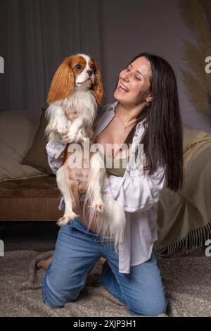 Junge schwangere Frau, die zu Hause auf dem Sofa sitzt. Er lacht fröhlich und spielt mit seinem Hund. Kavalierkönig charles, Cocker Spaniel Stockfoto