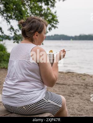 Eine übergewichtige Frau mittleren Alters ist draußen in der Nähe eines Sees und isst Nahrung aus einer Thermoskanne. Stockfoto