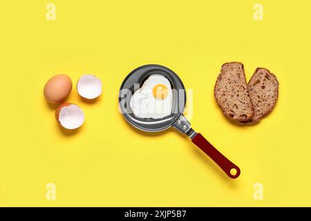 Eierschale, Stück Brot und herzförmiges Spiegelei in der Pfanne auf gelbem Hintergrund Stockfoto