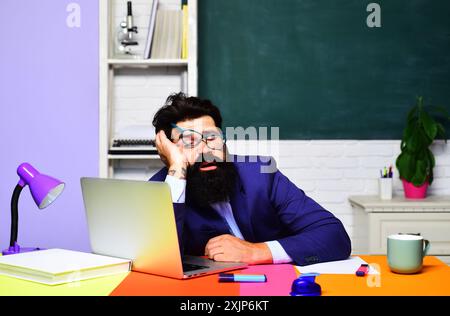 Müder Lehrer schläft nach dem Vortrag im Klassenzimmer. Schulferien. Ermüdeter männlicher Schullehrer, der auf dem Schreibtisch schläft. Überarbeiteter Hochschulstudent Stockfoto