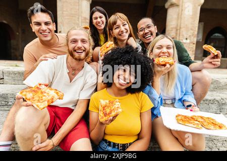 Junge Gruppe von verschiedenen Freunden lächelt in die Kamera, während sie draußen Pizza essen Stockfoto