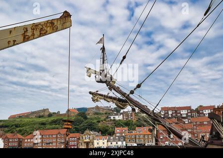 Blick auf einen historischen Schiffsmast und Takelage mit einer Küstenstadt und einem Hügel im Hintergrund. Der Himmel ist teilweise bewölkt. Stockfoto