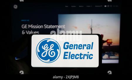 Stuttgart, Deutschland, 07.01.2023: Person mit Mobiltelefon mit Logo des amerikanischen Konzerns General Electric Company (GE) vor dem Bildschirm Stockfoto