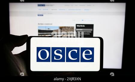 Stuttgart, Deutschland, 28.12.2023: Person, die ein Mobiltelefon mit dem Logo der Organisation für Sicherheit und Zusammenarbeit in Europa (OSZE) vor dem Internet hält Stockfoto
