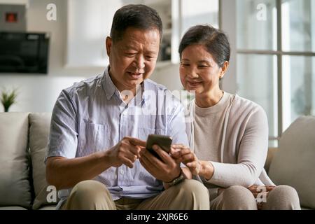 Glückliches älteres asiatisches Paar, das zu Hause auf der Couch sitzt und gemeinsam das Handy benutzt Stockfoto