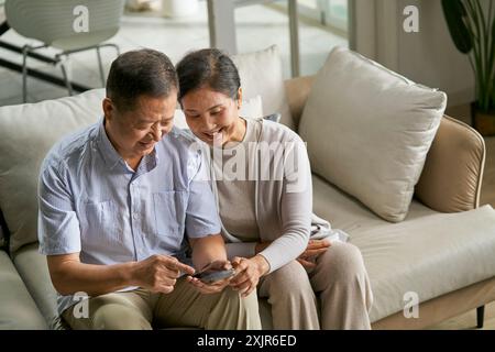 Glückliches älteres asiatisches Paar, das zu Hause auf der Couch sitzt und gemeinsam das Handy benutzt Stockfoto