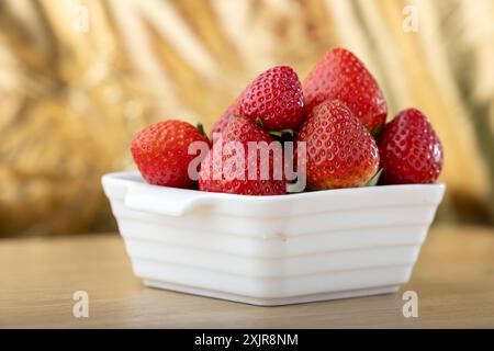 Frische, reife Erdbeeren glitzern in einer weißen, strukturierten Schüssel auf einer Holzoberfläche. Die Erdbeeren sind leuchtend rot und zeigen ihren saftigen Reiz Stockfoto