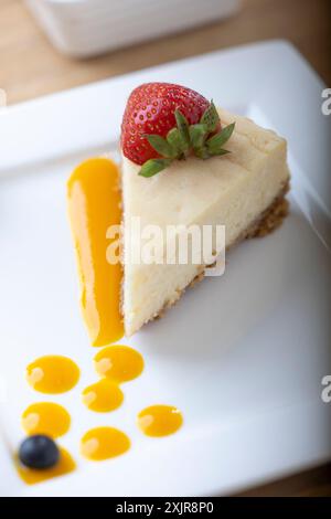 Eine Scheibe cremiger Käsekuchen mit einer frischen Erdbeere, serviert auf einem weißen Teller mit einem Hauch Fruchtsauce. Eine Schüssel mit frischen Erdbeeren Stockfoto