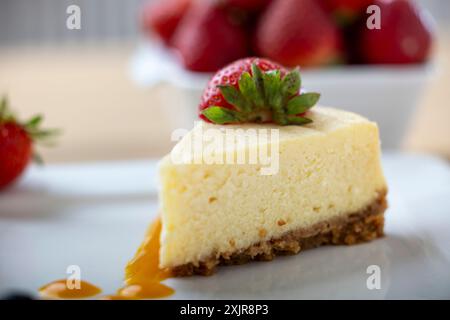 Eine Scheibe cremiger Käsekuchen mit einer frischen Erdbeere, serviert auf einem weißen Teller mit einem Hauch Fruchtsauce. Eine Schüssel mit frischen Erdbeeren Stockfoto