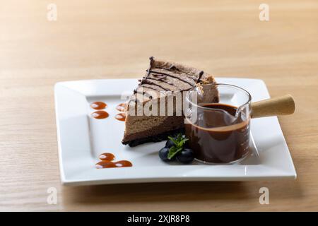 Dekadente Scheibe Schokoladen-Käsekuchen mit Schokoladensoße, serviert auf einem weißen quadratischen Teller. Begleitet von einer kleinen Tasse reichhaltigen Schokoladen-gan Stockfoto