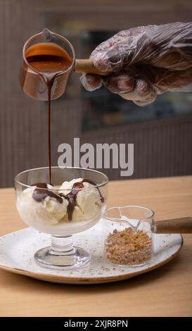 Eine Hand, die einen Plastikhandschuh trägt, gießt eine reiche Schokoladensauce über einen Messlöffel Vanilleeis in eine Glasschale, die auf eine weiße Platte gelegt wird. Stockfoto