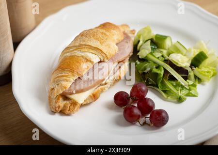 Ein frisch gebackenes Croissant-Sandwich gefüllt mit Schinken und Käse, serviert auf einem weißen Teller mit gemischtem grünem Salat und ein paar roten Trauben. Stockfoto