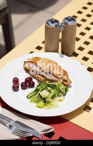 Ein frisch gebackenes Croissant-Sandwich gefüllt mit Schinken und Käse, serviert auf einem weißen Teller mit gemischtem grünem Salat und ein paar roten Trauben. Stockfoto