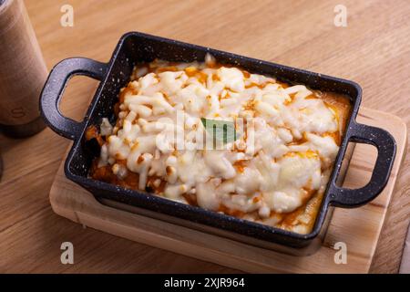 Eine köstliche, käsige Lasagne, serviert in einem schwarzen Keramikgericht, garniert mit geschmolzenem Mozzarella und garniert mit einem Basilikumblatt. Stockfoto