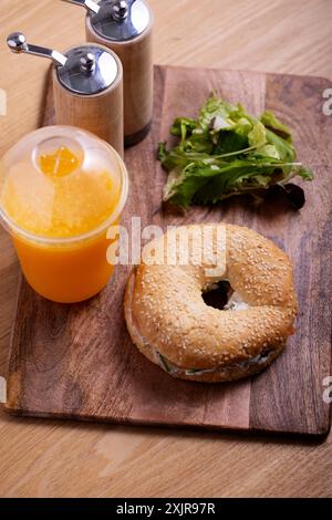 Leckerer Sesam-Bagel mit frischem Frischkäse und Gemüse auf einem Holzbrett serviert, gepaart mit einem erfrischenden Orangensaft in einer Plastiktasse. Stockfoto