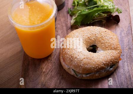 Leckerer Sesam-Bagel mit frischem Frischkäse und Gemüse auf einem Holzbrett serviert, gepaart mit einem erfrischenden Orangensaft in einer Plastiktasse. Stockfoto