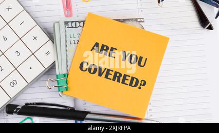 Sind Sie bedeckt auf einem gelben Aufkleber neben amerikanischen 100-Dollar-Scheinen, einem Taschenrechner, einem Stift auf weißem Hintergrund Stockfoto