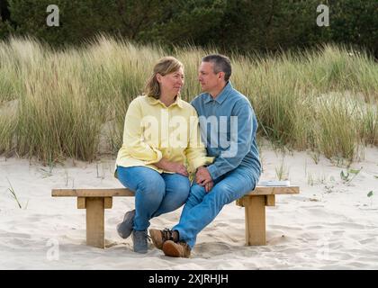 Ein Paar mittleren Alters im Urlaub auf dem Meer. Ein Mann und eine Frau genießen ihren Urlaub. Sie sitzen auf einer Bank an der Ostsee, mit Dünen in Backgr Stockfoto