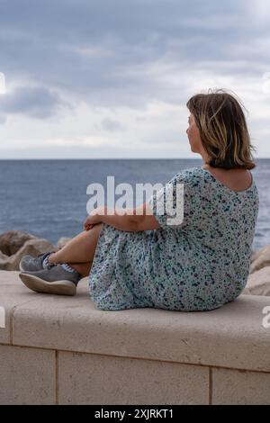 Eine Frau mittleren Alters sitzt am Strand und schaut in die Ferne. Genießt Leben, Frieden, Zufriedenheit. Meer und Berge am Horizont. Stockfoto