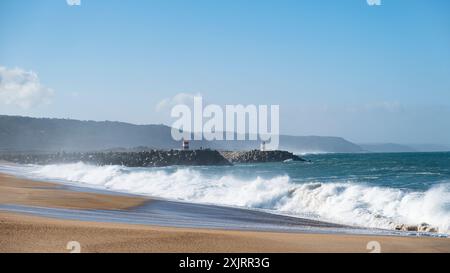 Wellen stürzen auf einen Sandstrand in der Nähe eines felsigen Wellenbrechers mit roten und weißen Leuchttürmen unter einem klaren blauen Himmel. Stockfoto