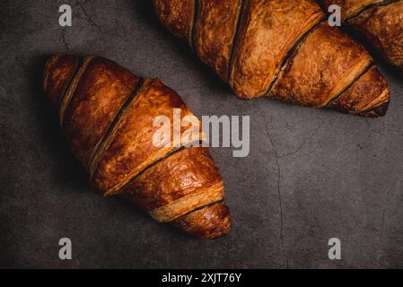 Drei frisch gebackene Croissants sitzen auf einer dunkelgrauen Arbeitsplatte, bereit zum Verzehr. Ihre goldbraune Kruste glitzert unter dem weichen Licht und lädt ein Stockfoto