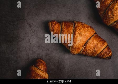 Drei frisch gebackene Croissants sitzen auf einer dunkelgrauen Arbeitsplatte, bereit zum Verzehr. Ihre goldbraune Kruste glitzert unter dem weichen Licht und lädt ein Stockfoto