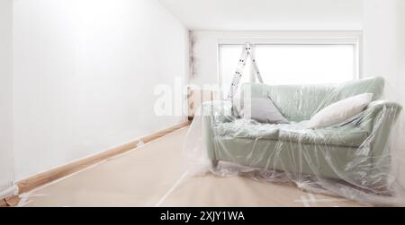 Leerer Wohnraum wird gerade renoviert, mit weißer Wand, Leiter und Sofa, die mit Plastiklaken von Malern bedeckt sind, vor dem hellen Fenster. Gussformreinigung und Wa Stockfoto