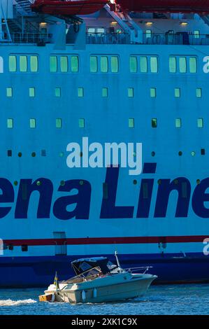 Ein kleines, weißes Motorboot mit dunkelblauem Baldachin wird vor einer großen Stena Line Fähre in einem ruhigen Hafen segelt. Die Fähre ist angedockt und ihr bl Stockfoto