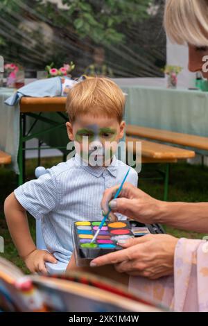 Das Mädchen, die Künstlerin hält Farben und zeichnet ein Bild auf das Gesicht eines kleinen Jungen. Das Gesicht eines kleinen Jungen mit einem Muster im Gesicht. Stockfoto