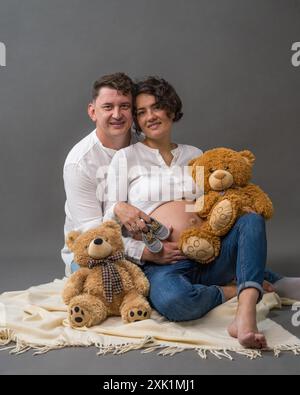 Eine glückliche schwangere Frau und ihr Mann sitzen auf dem Boden und halten zwei Teddybären in der Hand. Sie sehen sich mit Liebe an. Husban Stockfoto