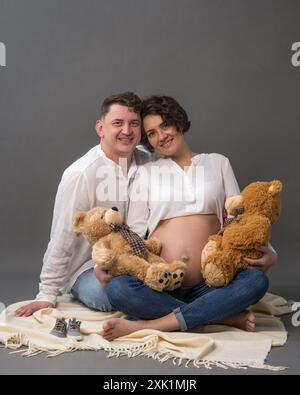 Eine glückliche schwangere Frau und ihr Mann sitzen auf dem Boden und halten zwei Teddybären in der Hand. Sie sehen sich mit Liebe an. Husban Stockfoto