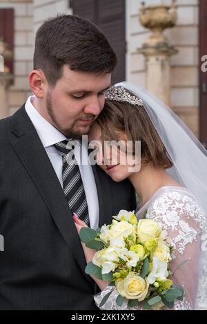 Hochzeitsporträt eines jungen, schönen, modernen Paares. Der Bräutigam umarmt die Braut, sie klammerte sich an ihn und hielt einen Strauß weißer Rosen. Berühren, Liebe, Fühlen Stockfoto