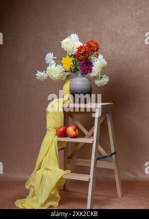 Viele helle, bunte Blumen auf der Holztreppe. Gelber Schal. Unten sind zwei rote Äpfel. Orangefarbener Hintergrund. Stillleben. Weißes, gelbes, rotes Aster Stockfoto