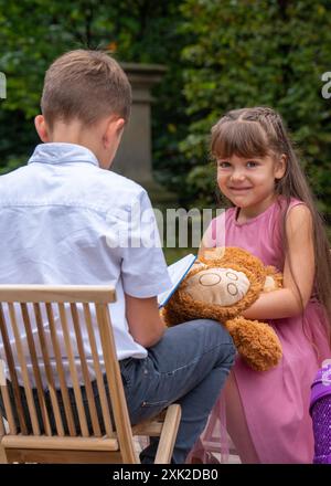 Der Junge sitzt mit dem Rücken auf einem Stuhl und liest ein Buch. Das Mädchen schaut mit Interesse rein und will sich die Bilder ansehen. Mädchen, 6 Jahre alt, mit einer Stockfoto