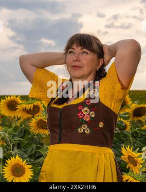 Eine Frau steht auf einem Feld mit gelben Sonnenblumen. Ihr Haar ist geflochten, sie trägt eine gelbe Bluse, ein braunes Sommerkleid und eine gelbe Schürze. Stockfoto