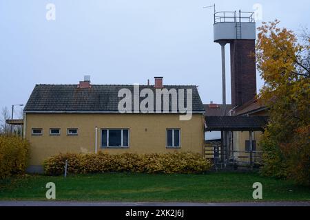 Ein einfaches, gelbes einstöckiges Gebäude mit einem Ziegeldach und kleinen rechteckigen Fenstern, das sich vor einem bewölkten Himmel befindet. Das Gebäude wird vom Herbst flankiert Stockfoto