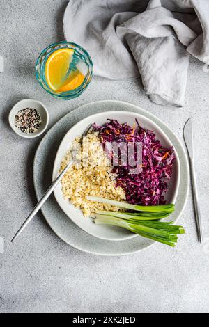 Ein nahrhaftes und lebhaftes Mittagessen mit rohen Rotkohl- und Karottensalat sowie gedämpftem Bulgur, serviert mit grüner Zwiebeln und Zitruswasser Stockfoto