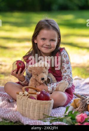 Ein kleines Mädchen sitzt auf einer Decke und hält einen Teddybären. Lächelt und schaut in die Kamera. In der Nähe liegt ein Korb mit roten Äpfeln. Picknick im Stockfoto