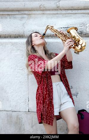 Eine junge Frau spielt leidenschaftlich Saxophon auf einer Stadtstraße, steht vor einer architektonischen Steinmauer, gekleidet in einem roten Hemd und einem Stockfoto